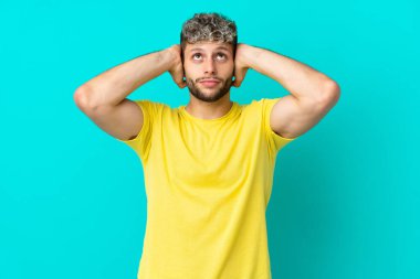 Mavi arka planda izole edilmiş yakışıklı beyaz adam hayal kırıklığına uğramış ve kulaklarını kapatmış.