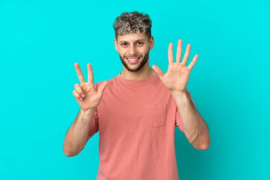 Mavi arka planda izole edilmiş yakışıklı beyaz adam parmaklarıyla sekizi sayıyor.