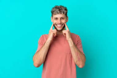 Mavi arka planda izole edilmiş yakışıklı beyaz adam mutlu ve hoş bir ifadeyle gülümsüyor.