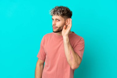 Genç beyaz adam mavi arka planda izole edilmiş bir şekilde elini kulağına koyarak bir şeyler dinliyor.