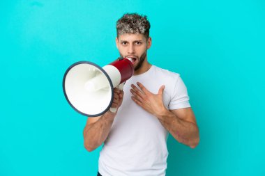 Young handsome caucasian man isolated on blue background shouting through a megaphone with surprised expression