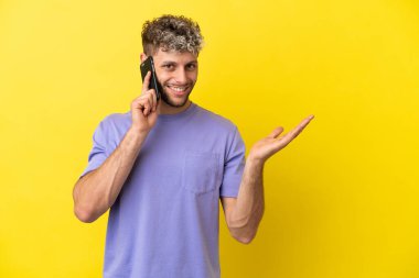 Young caucasian man using mobile phone isolated on yellow background extending hands to the side for inviting to come