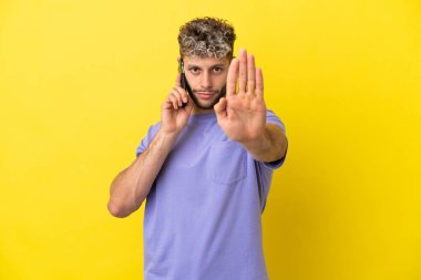 Sarı arka planda izole edilmiş cep telefonu kullanan beyaz bir adam dur hareketi yapıyor.