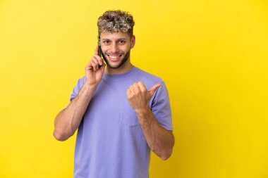 Sarı arka planda izole edilmiş cep telefonu kullanan genç beyaz adam bir ürünü sunmak için yan tarafı işaret ediyor.