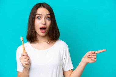 Dişlerini fırçalayan genç Ukraynalı kadın mavi arka planda izole edilmiş şaşırmış ve parmağını yana doğru işaret ediyor.