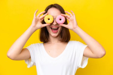 Young Ukrainian woman isolated on yellow background holding donuts in an eye