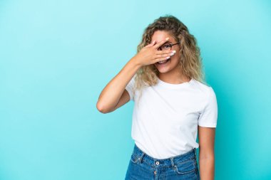 Kıvırcık saçlı kız mavi arka planda izole edilmiş gözleri elle kaplanmış ve gülümsüyor.