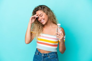 Mavi arka planda izole edilmiş bir şişe suyu olan genç sarışın kadın gülüyor.