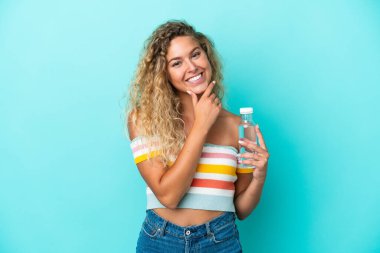 Mavi arka planda bir şişe suyu olan genç sarışın kadın mutlu ve gülümsüyor.