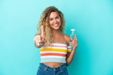 Mavi arka planda su şişesi olan genç sarışın kadın parmağını kaldırıyor ve gösteriyor.