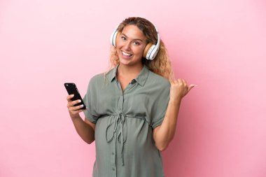 Kıvırcık saçlı kız pembe arka planda izole edilmiş hamile ve işaret ederken müzik dinliyor.