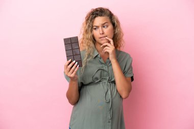Kıvırcık saçlı kız pembe arka planda izole edilmiş hamile ve elinde çikolata tutarken şüpheleri var.