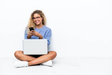 Dizüstü bilgisayarı olan kıvırcık saçlı kız yerde oturuyor ve cep telefonuyla mesaj yolluyor.