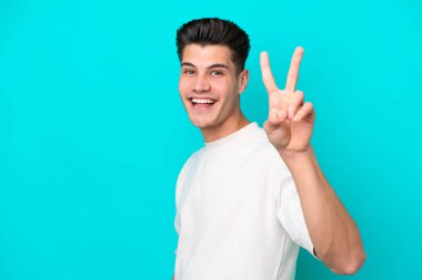 Genç, yakışıklı, beyaz bir adam mavi bir bakkalda izole edilmiş gülümsüyor ve zafer işareti gösteriyor.