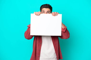 Genç, beyaz bir adam mavi bir bakkalda izole edilmiş boş bir levha tutuyor ve arkasında saklanıyor.