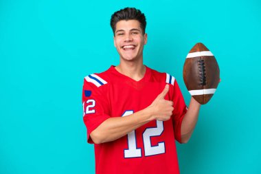 Rugby oynayan genç beyaz adam mavi arka planda izole edilmiş bir şekilde baş parmak hareketi yapıyor.