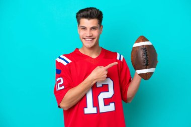 Rugby oynayan beyaz bir genç, mavi arka planda izole edilmiş bir şekilde bir ürünü sunmak için yan tarafa bakıyor.