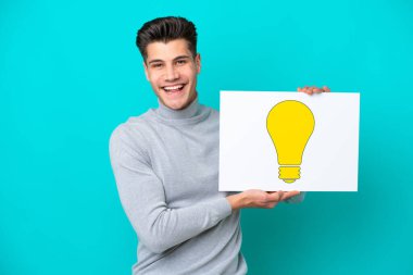 Genç, beyaz bir adam mavi bir bakkal sahasında izole edilmiş elinde mutlu ifadeli bir ampul simgesi olan bir pankart tutuyor.