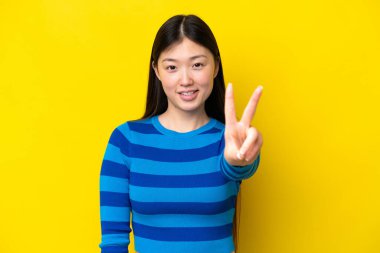 Sarı arka planda izole edilmiş genç Çinli kadın gülümsüyor ve zafer işareti gösteriyor.