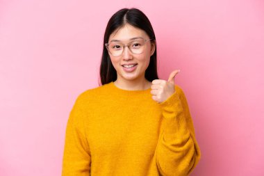 Genç Çinli bir kadın pembe arka planda gözlüklü ve başparmağı yukarıda.