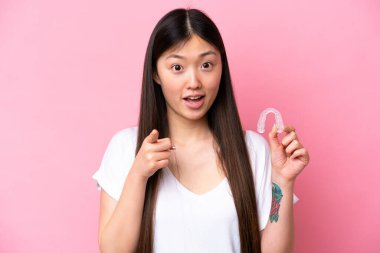 Pembe arka planda izole edilmiş görünmez diş telleri tutan genç Çinli kadın şaşırmış ve ön tarafı işaret ediyor.