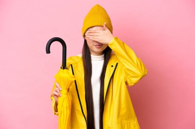 Young Chinese woman with rainproof coat and umbrella isolated on pink background covering eyes by hands. Do not want to see something