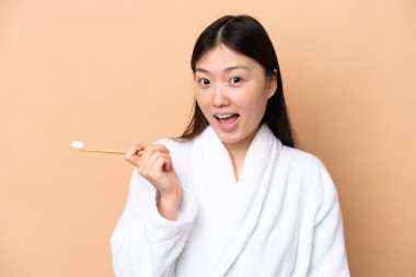 Genç Çinli kadın bej arka planda diş fırçası ve şaşırmış bir ifadeyle izole edilmiş.