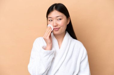 Yüzündeki makyajı silmek için bej arka planda pamuk pedi olan genç Çinli bir kadın.