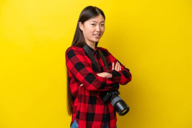 Sarı arka planda izole edilmiş genç fotoğrafçı kadın. Kolları çapraz ve ileriye bakıyor.