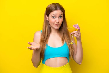Sarı arka planda izole edilmiş bir kokteyl tutan genç beyaz kadın omuzlarını kaldırırken şüpheyle hareket ediyor.