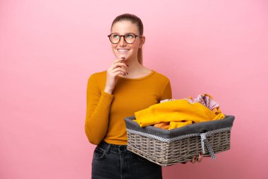 Pembe arka planda izole edilmiş çamaşır sepetini tutan beyaz bir kadın gülümserken yukarı bakıyor.