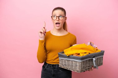 Pembe arka planda izole edilmiş çamaşır sepetini tutan beyaz bir kadın parmak kaldırmayı düşünüyor.