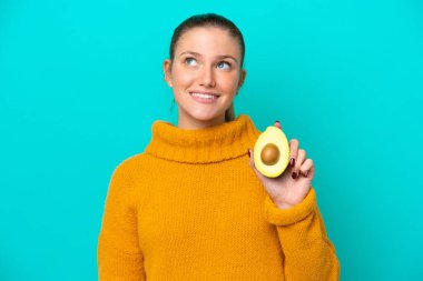 Avokado tutan beyaz bir kadın mavi arka planda izole edilmiş gülümserken yukarı bakıyor.