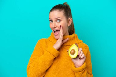 Avokado tutan beyaz bir kadın mavi arka planda izole edilmiş bir şekilde bir şeyler fısıldıyor.