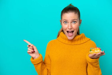 Mavi arka planda izole edilmiş tart tutan genç beyaz kadın şaşırdı ve parmağını yana çevirdi.