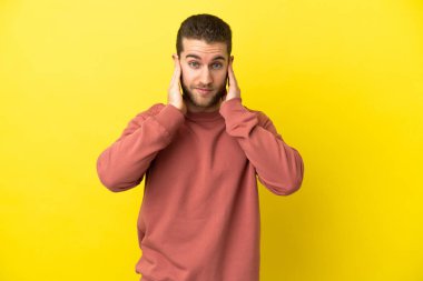 Sarışın yakışıklı adam izole edilmiş sarı arka planda hayal kırıklığına uğramış ve kulaklarını kapatmış.