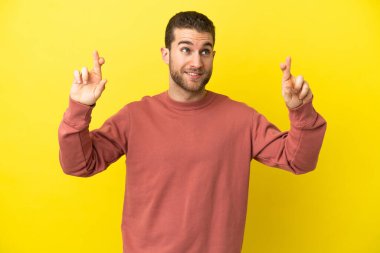 Sarışın yakışıklı adam izole edilmiş sarı arka planda parmaklarını çapraz yapıp en iyisini diliyor.