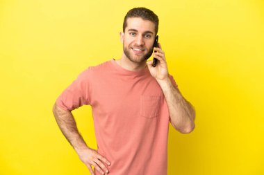 Yakışıklı sarışın adam izole edilmiş arka planda cep telefonu kullanıyor. Kolları kalçasında ve gülümsüyor.