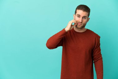 Yakışıklı sarışın adam, izole edilmiş mavi arka planda sessizlik işareti gösteriyor.