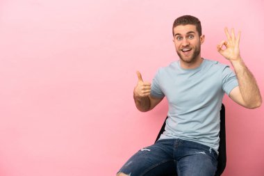 Young man sitting on a chair over isolated pink background showing ok sign and thumb up gesture