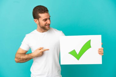 İzole edilmiş mavi arka planı olan yakışıklı sarışın. Üzerinde yeşil işaret simgesi olan bir levha tutuyor ve işaret ediyor.