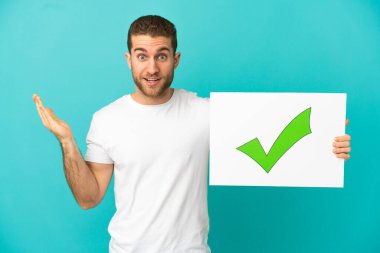 İzole edilmiş mavi arkaplanı olan yakışıklı sarışın. Üzerinde yeşil işaret simgesi olan bir pankart tutuyor.