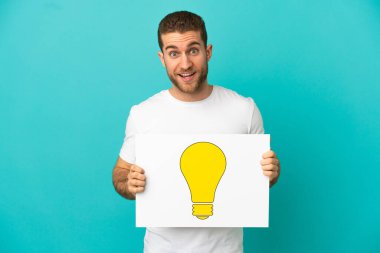 Yakışıklı, mavi arka planda, üzerinde mutlu yüz ifadesi olan ampul simgesi olan bir pankart tutan sarışın adam.