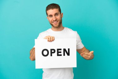 Yakışıklı, mavi arka planda, üzerinde açık yazı olan bir pankart tutan ve onu işaret eden bir sarışın.