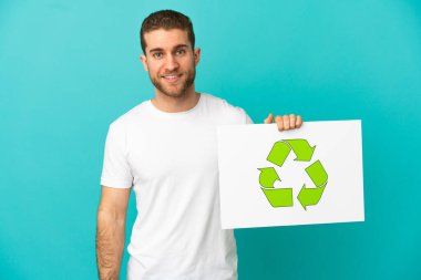 İzole edilmiş mavi arka planı olan yakışıklı sarışın adam elinde mutlu ifadeli geri dönüşüm simgesi olan bir pankart tutuyor.