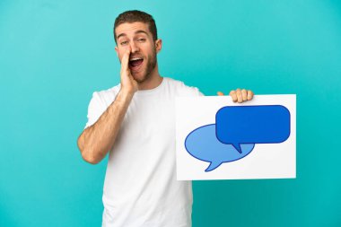 Yakışıklı sarışın, izole edilmiş mavi arkaplan üzerinde konuşma balonu ikonu olan bir levha tutuyor ve bağırıyor.
