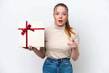 Beyaz arka planda izole edilmiş bir hediye tutan beyaz bir kadın şaşırmış ve ön tarafı işaret ediyor.