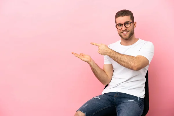 Jovem Sentado Uma Cadeira Sobre Fundo Rosa Isolado Segurando Copyspace — Fotografia de Stock