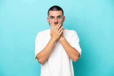 Mavi arka planda izole edilmiş genç beyaz adam elleriyle ağzını kapatıyor.