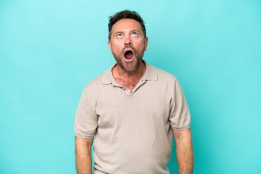 Orta yaşlı beyaz bir adam mavi arka planda izole edilmiş yukarı bakıyor ve şaşırmış bir ifadeyle